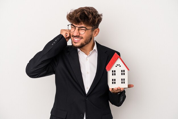 Homem jovem árabe imobiliário segurando uma casa modelo isolada no fundo isolado, cobrindo as orelhas com as mãos.