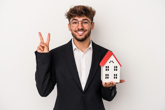 Homem jovem árabe imobiliário segurando uma casa modelo isolada no fundo isolado, mostrando o número dois com os dedos.