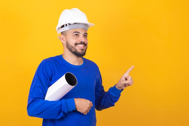 Homem jovem arquiteto com capacete e segurando plantas isoladas em fundo amarelo, apontando o dedo para o lado