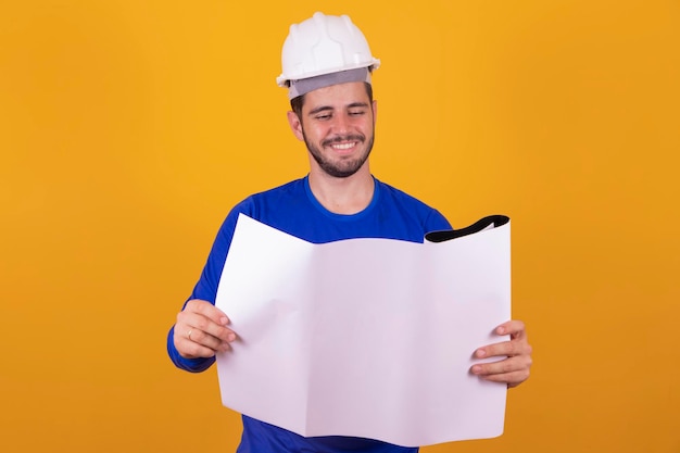 Homem jovem arquiteto com capacete e segurando plantas isoladas em fundo amarelo, apontando o dedo para o lado