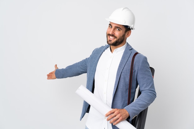 Homem jovem arquiteto com capacete e segurando plantas isoladas na parede branca, estendendo as mãos para o lado para convidar para vir