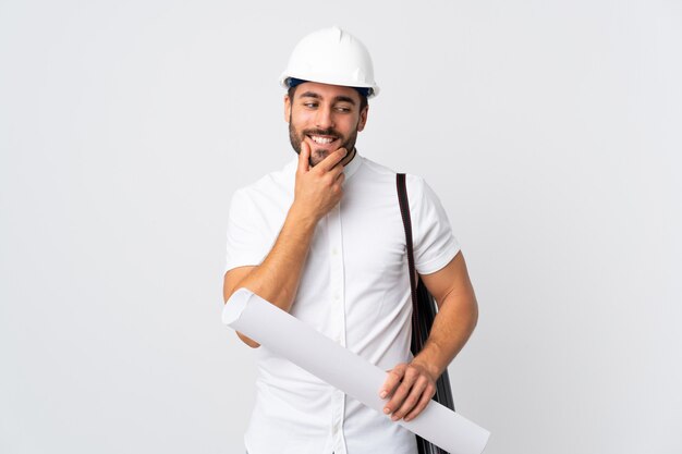 Homem jovem arquiteto com capacete e segurando plantas isoladas na parede branca, olhando para o lado e sorrindo