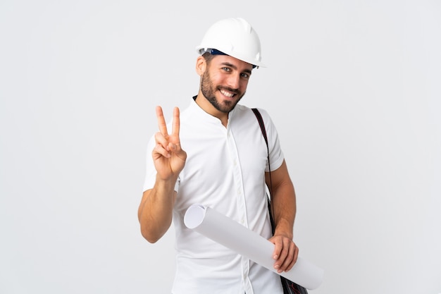 Homem jovem arquiteto com capacete e segurando plantas isoladas na parede branca, sorrindo e mostrando sinal de vitória