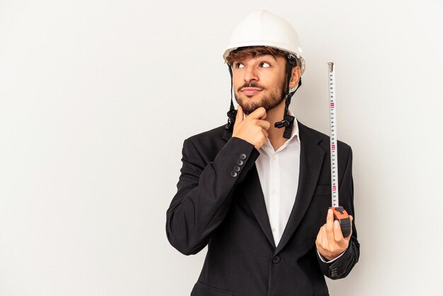 Homem jovem arquiteto de raça mista, usando um capacete e segurando o medidor isolado em um fundo cinza, olhando de soslaio com expressão duvidosa e cética.