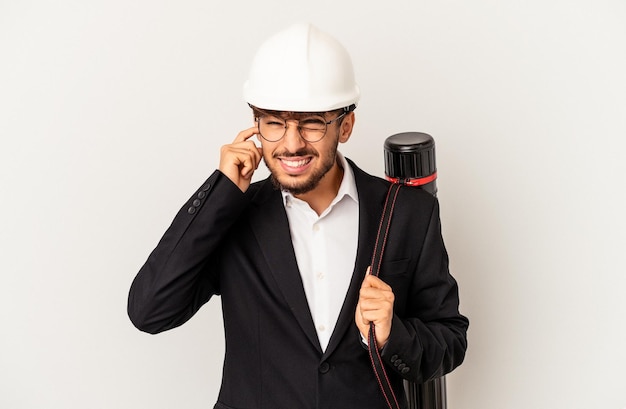 Homem jovem arquiteto de raça mista usando um capacete isolado em um fundo cinza, cobrindo as orelhas com as mãos.