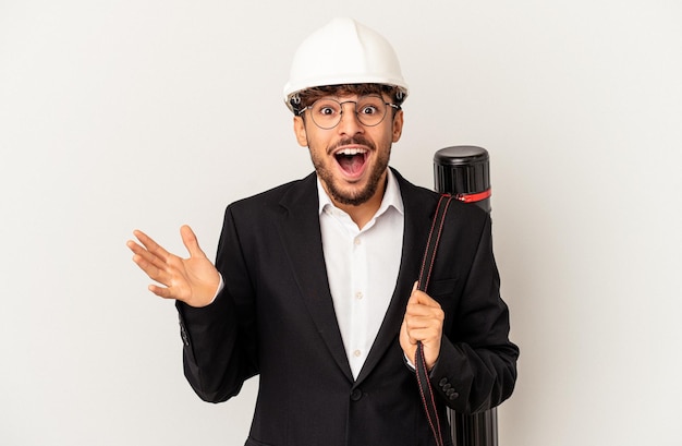 Homem jovem arquiteto de raça mista usando um capacete isolado no fundo cinza, recebendo uma agradável surpresa, animado e levantando as mãos.