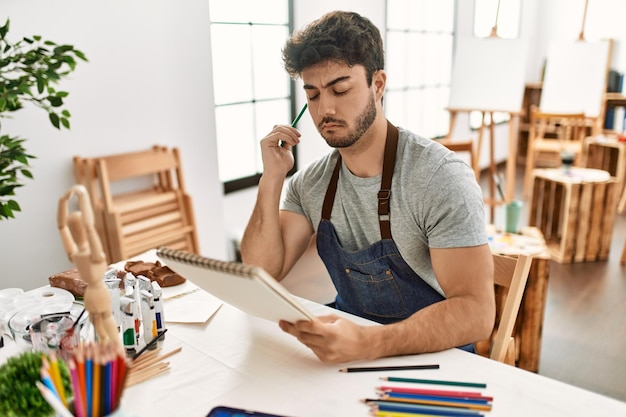 Homem jovem artista hispânico com pintura de expressão séria no estúdio de arte.