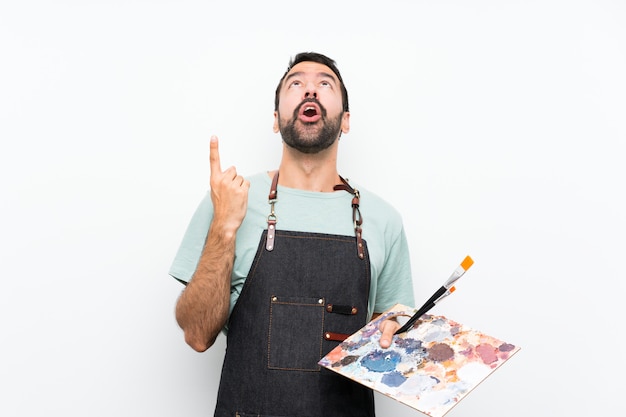 Foto homem jovem artista segurando uma paleta sobre parede isolada, apontando para cima e surpreso