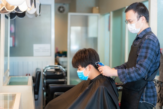 Homem jovem asiático e cabeleireiro usando máscara médica para se proteger durante o romance Coronavirus, Covid-19 no Barbershop Hair Care Service.