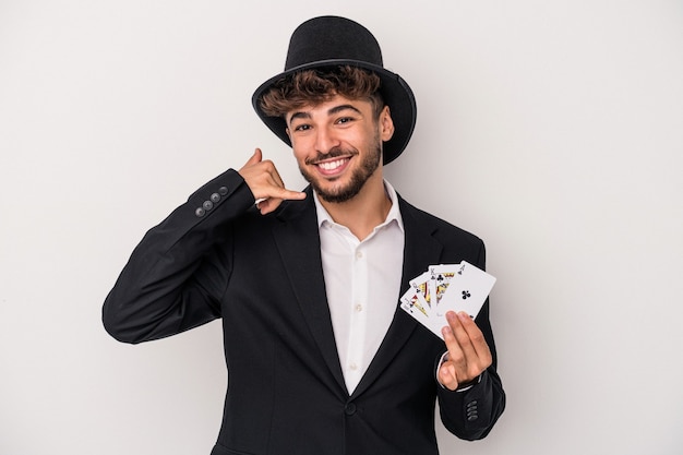 Homem jovem assistente árabe segurando cartas mágicas isoladas no fundo branco, mostrando um gesto de chamada de telefone móvel com os dedos.