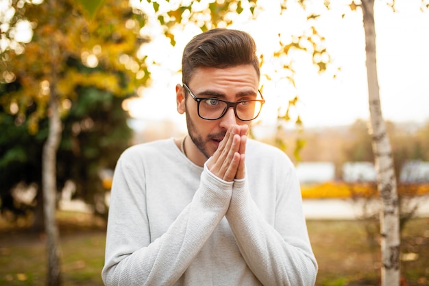 Homem jovem bonito hipster de óculos e uma camisola branca está andando em um parque de outono frio