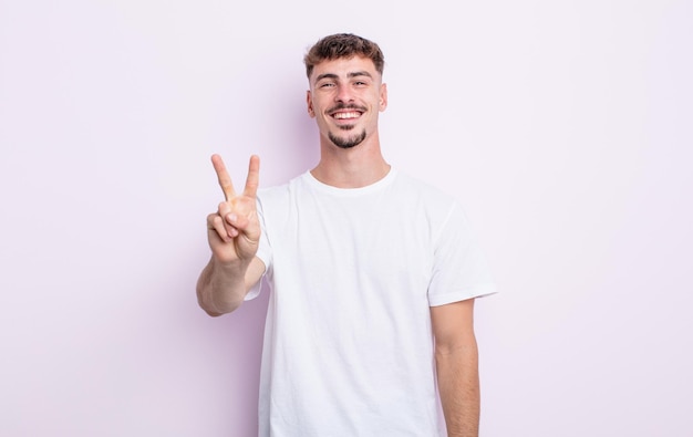 Homem jovem bonito sorrindo e parecendo amigável, mostrando o número dois ou o segundo com a mão para a frente, em contagem regressiva