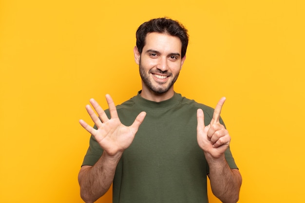 Homem jovem bonito sorrindo e parecendo amigável, mostrando o número sete ou sétimo com a mão para a frente, em contagem regressiva