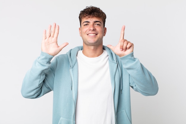 Homem jovem bonito sorrindo e parecendo amigável, mostrando o número sete ou sétimo com a mão para a frente, em contagem regressiva