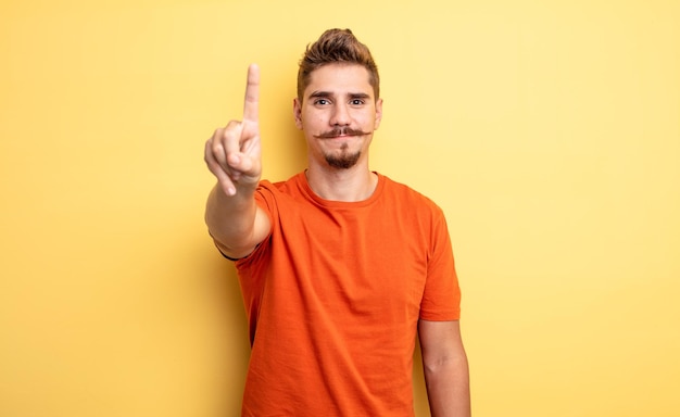 Homem jovem bonito sorrindo e parecendo amigável, mostrando o número um ou primeiro com a mão para a frente, em contagem regressiva. conceito de bigode estranho