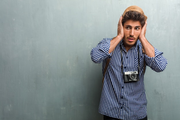 Homem jovem bonito viajante usando um chapéu de palha, uma mochila e uma câmera fotográfica cobrindo os ouvidos com as mãos, com raiva e cansado de ouvir algum som