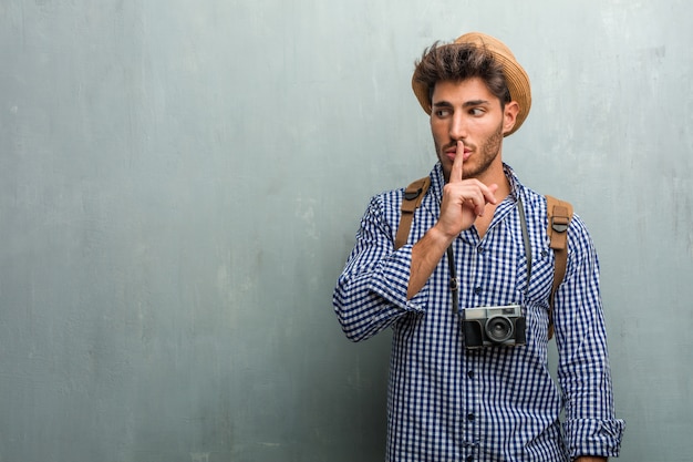 Homem jovem bonito viajante usando um chapéu de palha, uma mochila e uma câmera fotográfica mantendo um segredo ou pedindo silêncio, cara séria, conceito de obediência