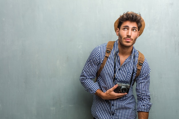 Homem jovem bonito viajante usando um chapéu de palha, uma mochila e uma câmera fotográfica, olhando para cima