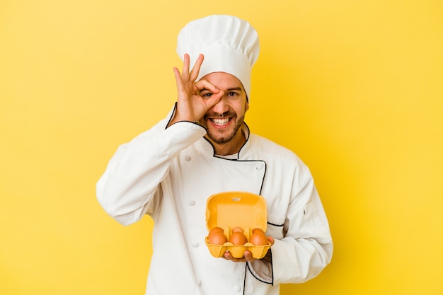 Homem jovem chef caucasiano segurando ovos isolados em fundo amarelo animado, mantendo o gesto ok no olho.