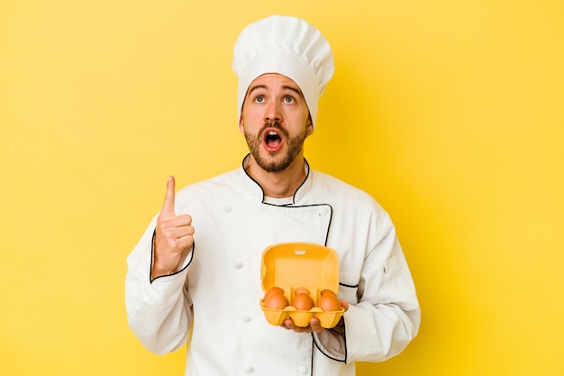 Homem jovem chef caucasiano segurando ovos isolados em um fundo amarelo, apontando para cima com a boca aberta.
