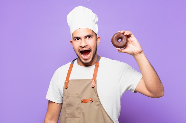 Homem jovem chef hispânico com expressão de surpresa segurando um donut