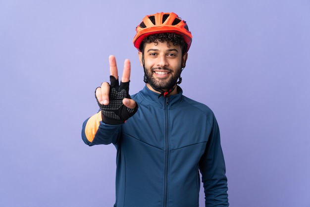 Homem jovem ciclista marroquino isolado na parede roxa sorrindo e mostrando sinal de vitória