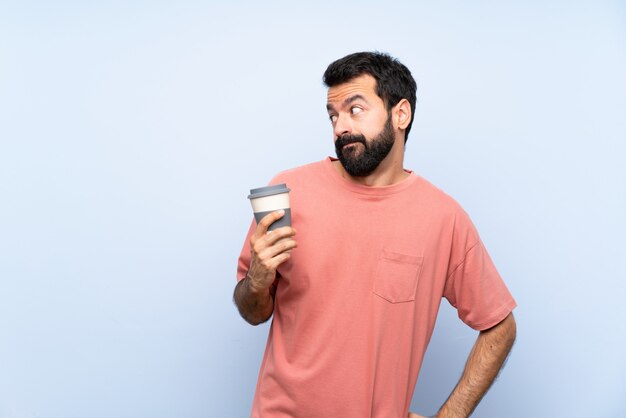 Homem jovem, com, barba, segurando, levar, café, isolado, azul, parede, fazendo, dúvida, gesto, olhar lado