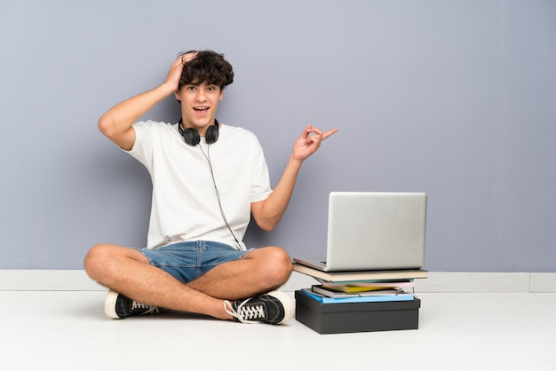 Homem jovem, com, seu, laptop, sentando, um, a, chão, surpreendido, e, apontar, dedo, ao lado