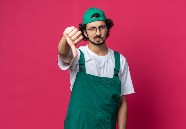 Homem jovem construtor carrancudo usando uniforme com boné mostrando o polegar para baixo