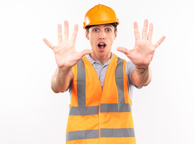 Homem jovem construtor com medo, de uniforme, mostrando gesto de parada isolado na parede branca