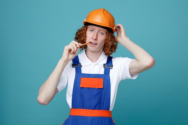 Homem jovem construtor insatisfeito de uniforme segurando lápis na boca isolado em fundo azul