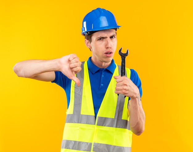 Homem jovem construtor insatisfeito, de uniforme, segurando uma chave de boca mostrando o polegar isolado na parede amarela