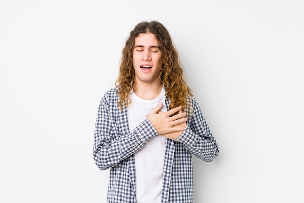 Homem jovem de cabelos longos posando isolado rindo mantendo as mãos no coração, o conceito de felicidade.