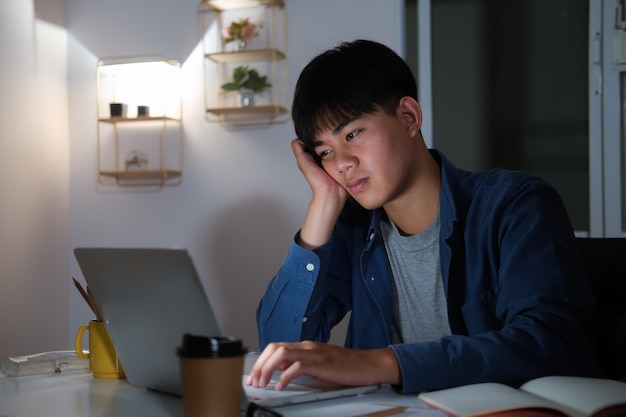 Homem jovem de colagem exausto com sono trabalhando horas extras na mesa do escritório com seu laptop, ele está prestes a adormecer, privação de sono e conceito de trabalho extra.