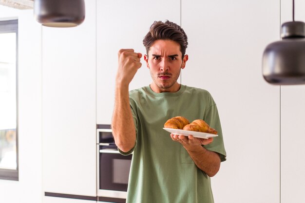 Homem jovem de raça mista comendo croissant em uma cozinha de manhã, mostrando o punho para a câmera, expressão facial agressiva.