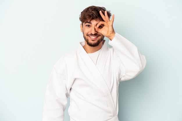 Homem jovem de raça mista fazendo caratê isolado no fundo azul animado, mantendo o gesto ok no olho.