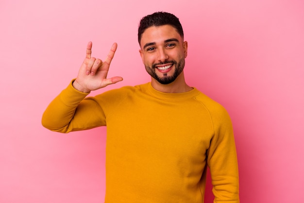 Homem jovem de raça mista isolado em fundo rosa, mostrando um gesto de chifres como um conceito de revolução.