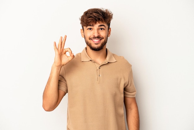 Homem jovem de raça mista isolado em um fundo cinza alegre e confiante, mostrando um gesto ok.