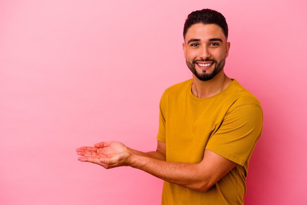 Homem jovem de raça mista isolado em um fundo rosa, segurando um espaço de cópia na palma da mão.
