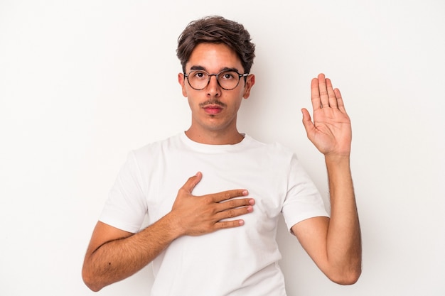 Homem jovem de raça mista, isolado no fundo branco, fazendo um juramento, colocando a mão no peito.