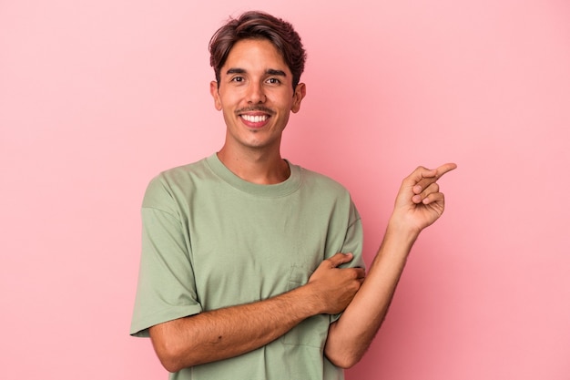 Homem jovem de raça mista isolado no fundo branco, sorrindo alegremente, apontando com o dedo indicador para longe.