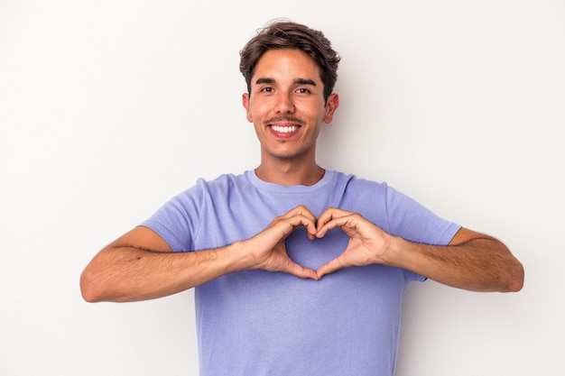 Homem jovem de raça mista isolado no fundo branco, sorrindo e mostrando uma forma de coração com as mãos.