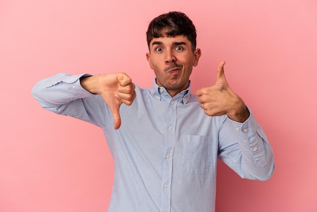 Homem jovem de raça mista isolado no fundo rosa, mostrando os polegares para cima e os polegares para baixo, difícil escolher o conceito