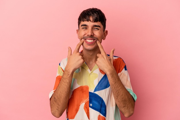 Homem jovem de raça mista isolado no fundo rosa sorri apontando os dedos para a boca