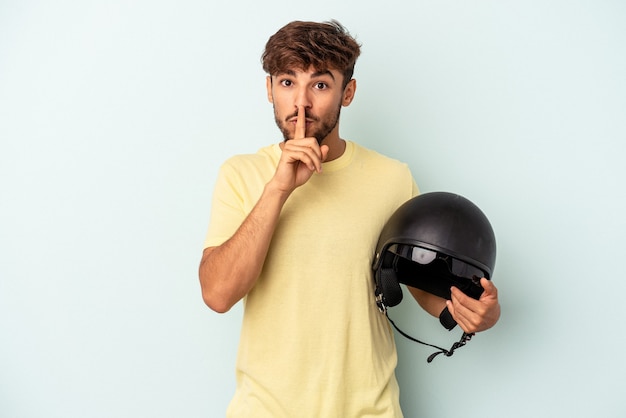 Homem jovem de raça mista segurando capacete de motocicleta isolado em fundo azul, mantendo um segredo ou pedindo silêncio.