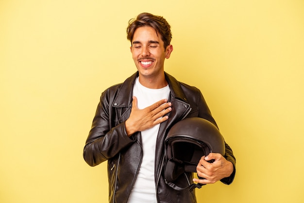 Homem jovem de raça mista segurando capacete isolado em fundo amarelo ri alto, mantendo a mão no peito.