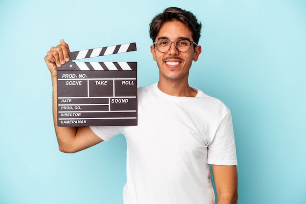 Homem jovem de raça mista segurando claquete isolada em fundo azul feliz, sorridente e alegre.