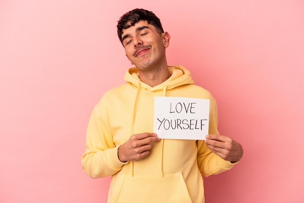 Homem jovem de raça mista, segurando o cartaz de amor a si mesmo isolado no fundo rosa