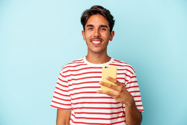 Homem jovem de raça mista, segurando o telefone móvel isolado no fundo azul, feliz, sorridente e alegre.