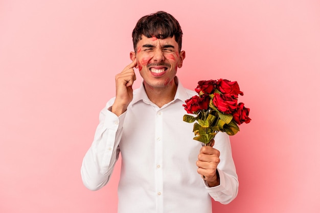 Homem jovem de raça mista, segurando um buquê de flores isoladas no fundo rosa, cobrindo os ouvidos com as mãos.